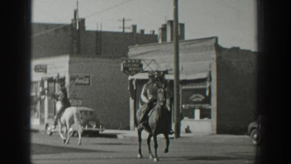 A dark horse and rider face toward the camera as another rider moves away past vintage cars and storefronts in Wyoming, 1939.