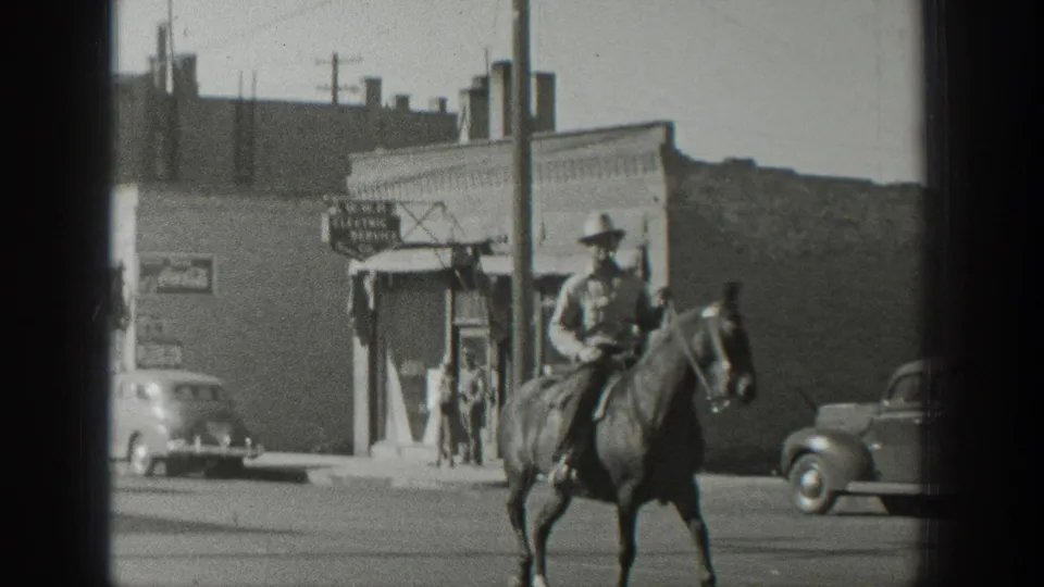 In 1939 Wyoming home-movie footage, a mounted man rides along a main street with storefronts, parked cars, and a signboard behind him.
