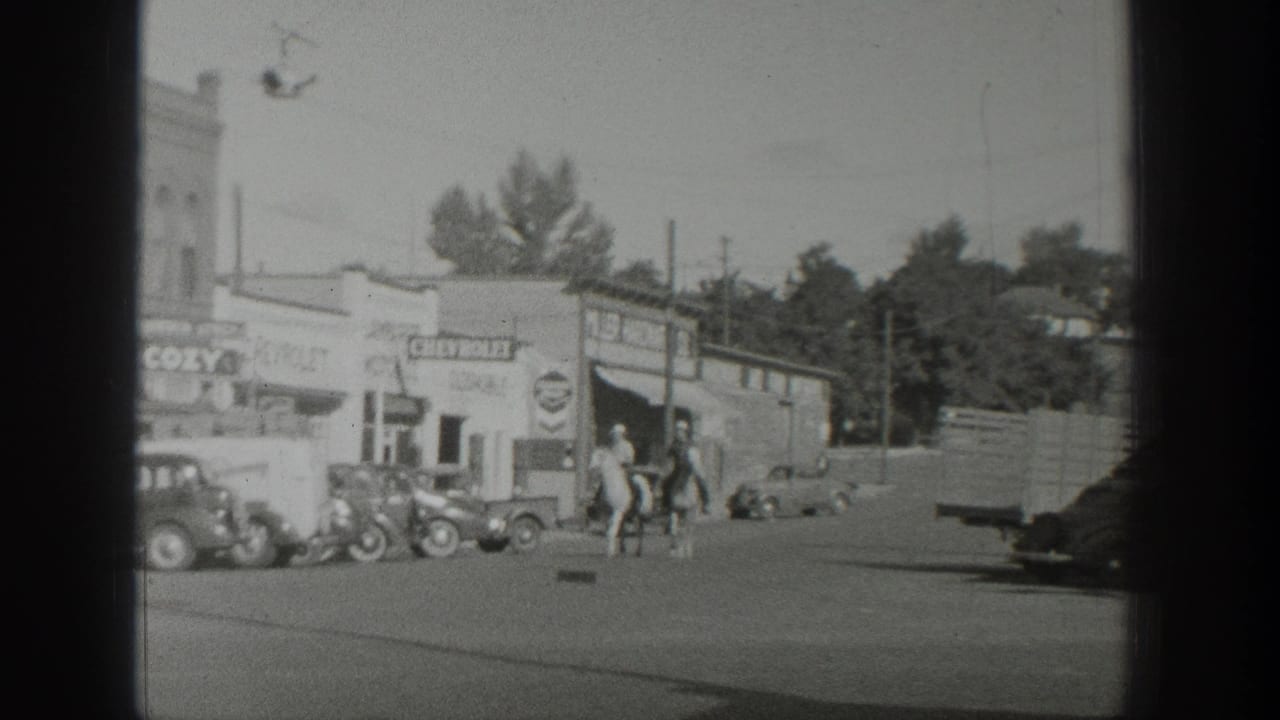 Several riders on horses pass storefronts with parked cars along the street.