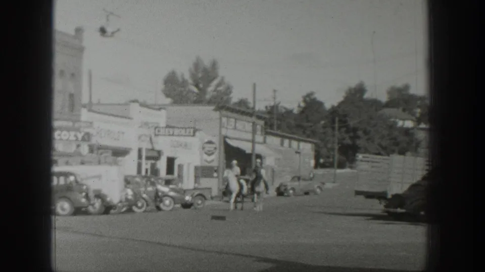 Three mounted men ride through a Wyoming town street in 1939, with Chevrolet and COZY signage and several 1930s cars visible nearby.
