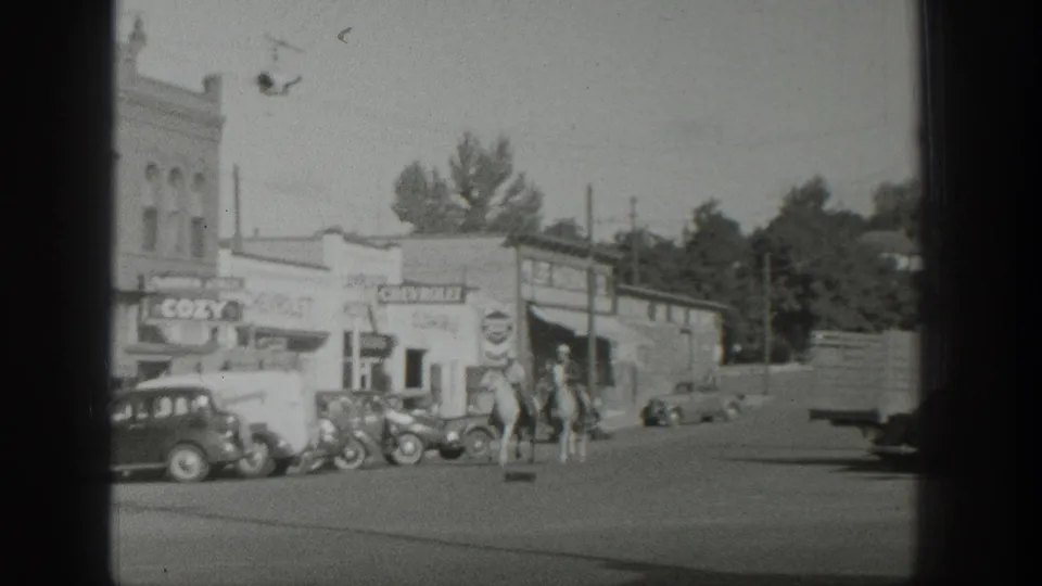 The riders move along the bright main street, framed by 1930s parked cars, storefront signage, utility poles, and trees in the background.