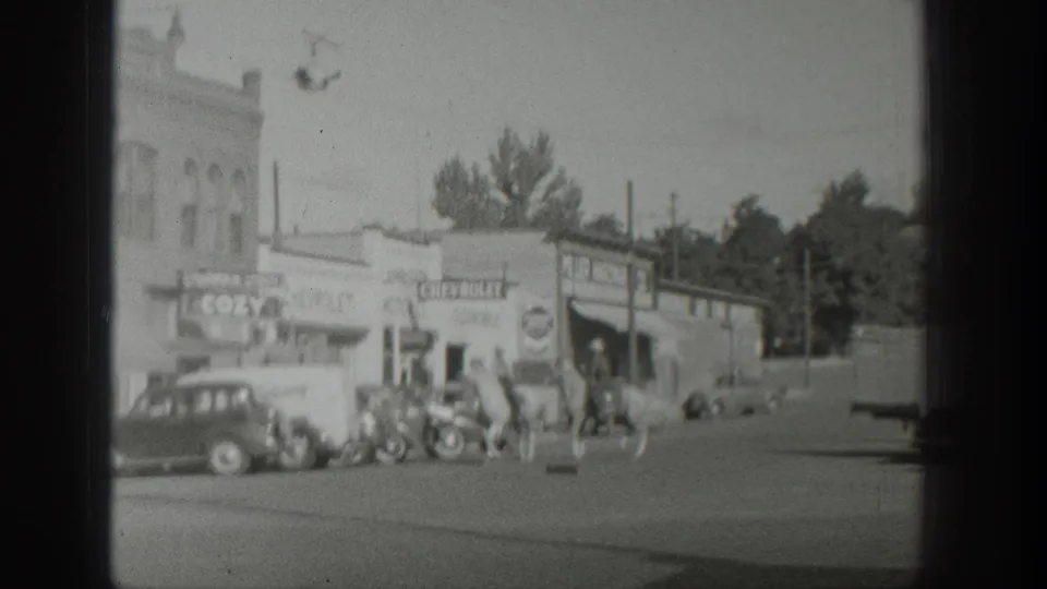 At the end of the clip, mounted men ride farther along the Wyoming town street, with storefront signs, automobiles, and trees still visible.