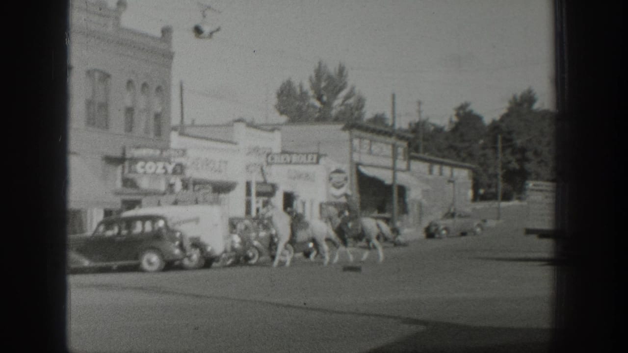 In 1939 Wyoming home-movie footage, mounted riders move past storefronts, a COZY sign, and vintage automobiles on a commercial street.