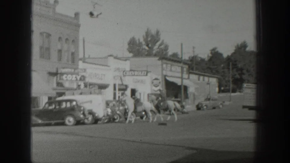 In 1939 Wyoming home-movie footage, mounted riders move past storefronts, a COZY sign, and vintage automobiles on a commercial street.