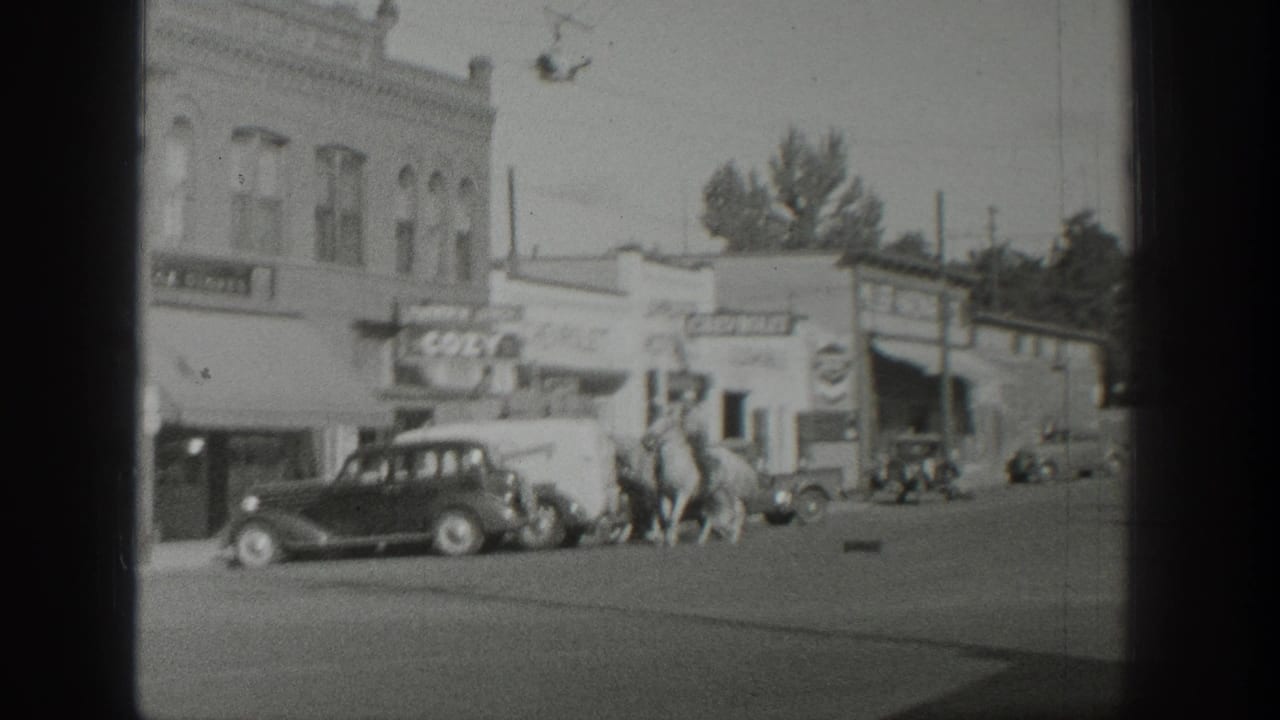 Two horseback riders move along a street lined with cars and storefront signage.