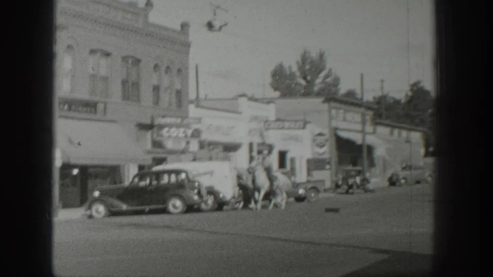 Two riders on horseback travel along a Wyoming town street in 1939, with cars, sidewalks, storefronts, and visible commercial signs nearby.