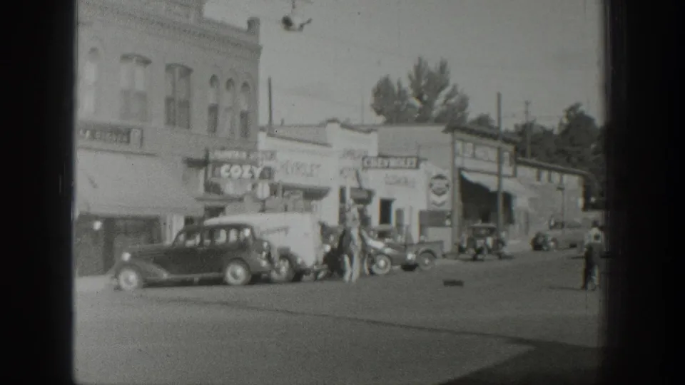 The riders continue through a 1939 Wyoming commercial street, passing automobiles, a COZY storefront sign, and low brick buildings.