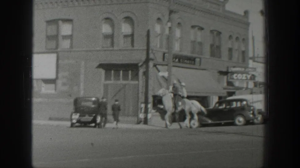 At the end of the shot, horseback riders pass a brick corner building and parked cars on a Wyoming town street in 1939.