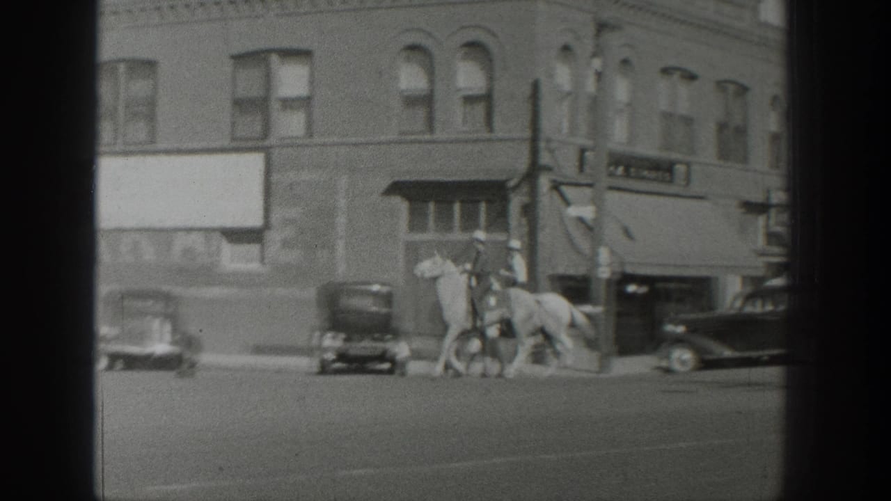 1939 Wyoming home-movie frame showing riders on horseback near parked cars and a brick storefront on a broad street.