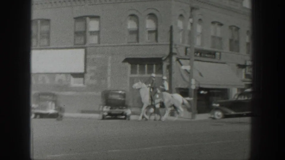 1939 Wyoming home-movie frame showing riders on horseback near parked cars and a brick storefront on a broad street.