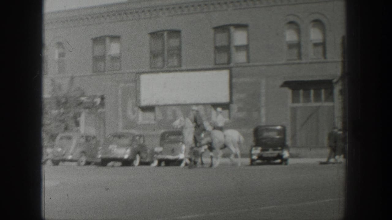 Two mounted riders are seen near parked cars in front of a brick building.