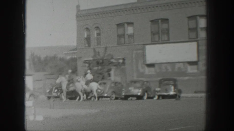 Late frame from 1939 Wyoming footage showing mounted riders moving past parked cars near a brick commercial building.
