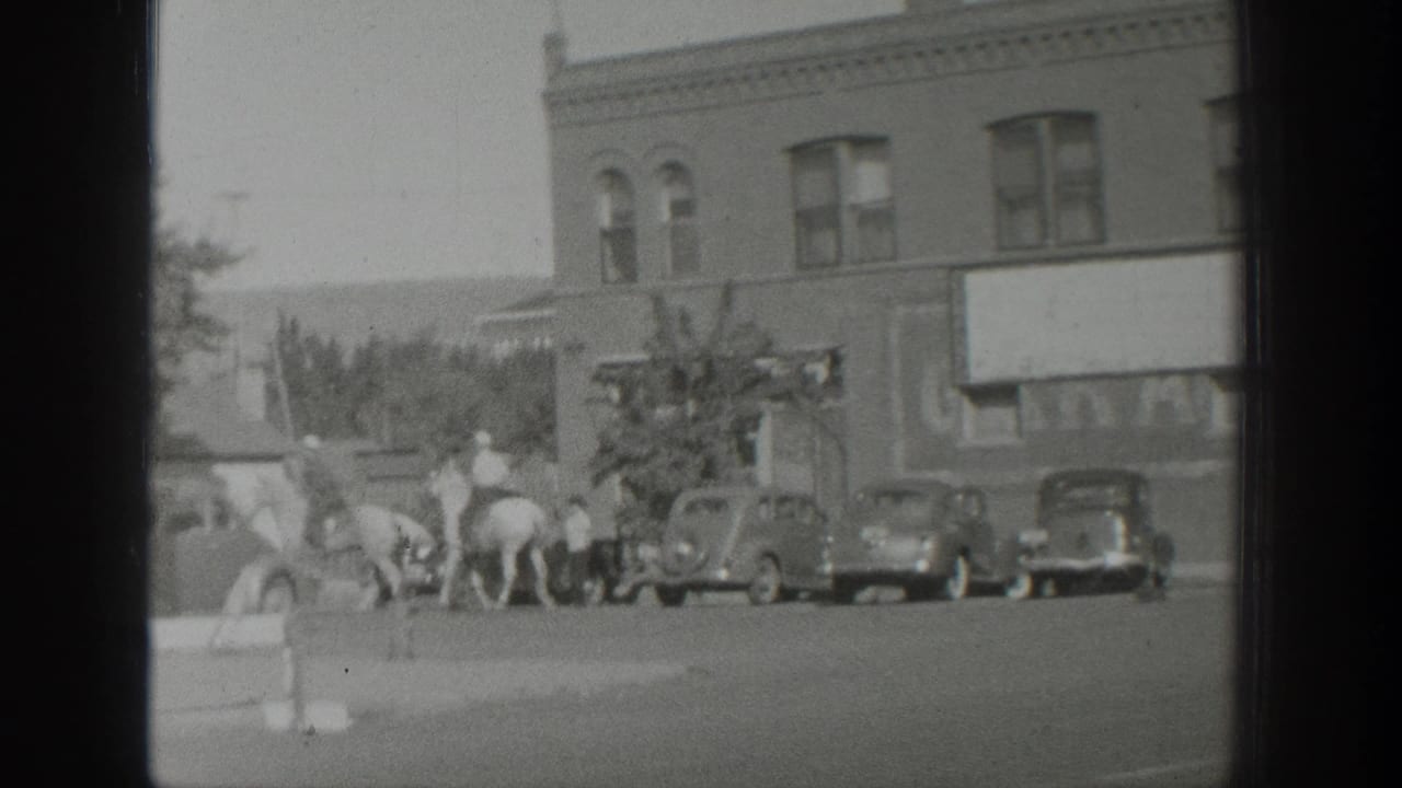1939 Wyoming home-movie frame showing riders on horseback crossing a small-town street near parked automobiles and brick storefronts.