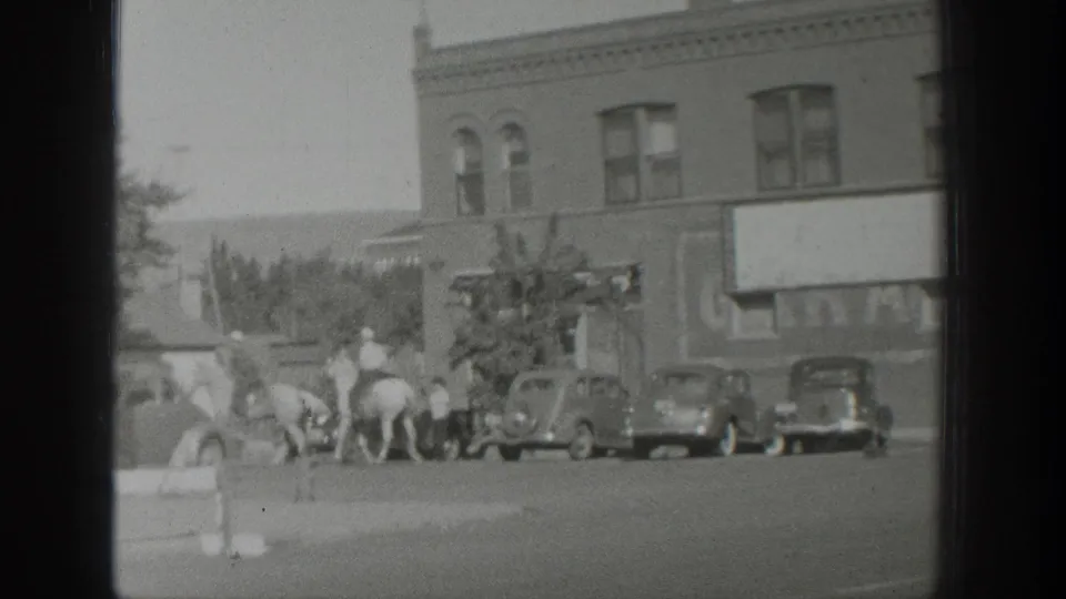 1939 Wyoming home-movie frame showing riders on horseback crossing a small-town street near parked automobiles and brick storefronts.