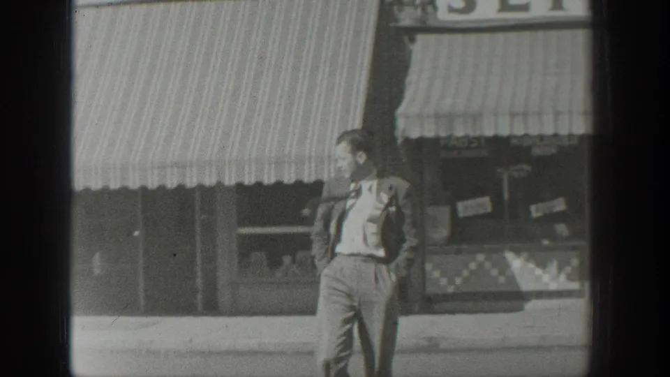 A suited man stands on a Wyoming sidewalk in front of striped storefront awnings and display windows in 1939 home-movie footage.