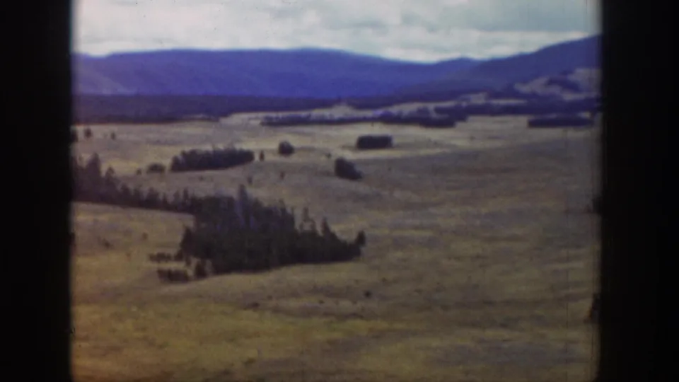 Color 8mm frame of a Wyoming valley panorama, with pale fields, dark tree clusters, distant ridges, and a cloudy mountain horizon.