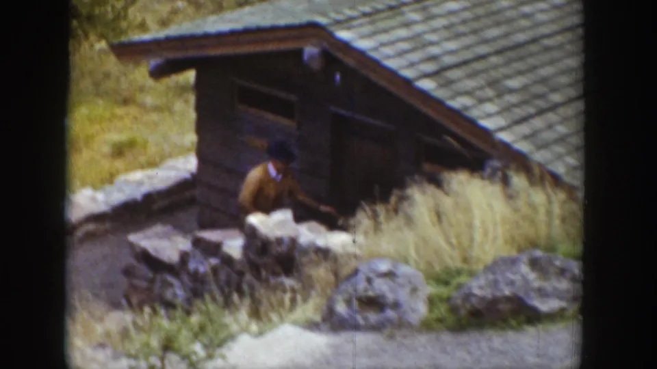 1939 Wyoming 8mm frame showing a rustic wooden cabin in a rocky setting, with an adult man partly visible near the doorway or path.
