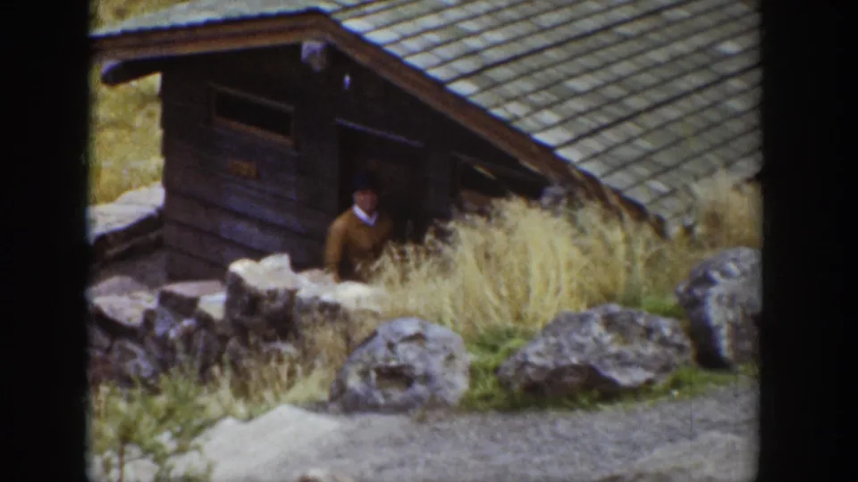 1939 Wyoming 8mm frame with an adult man by a rustic cabin entrance, looking out across rocks, dry grass, and a rugged outdoor setting.