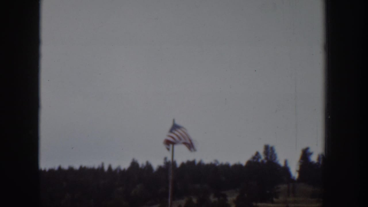 1939 Wyoming home-movie view of an American flag flying above a dark tree line under a cloudy sky.