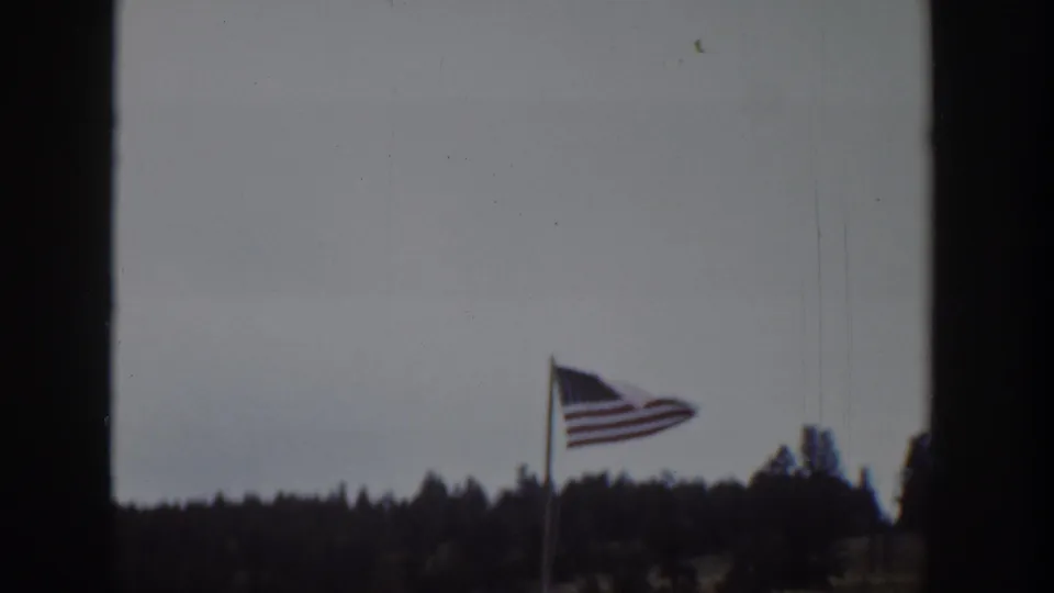 1939 Wyoming 8mm frame of a windblown American flag with a pale sky, forested ridge, and soft film grain.