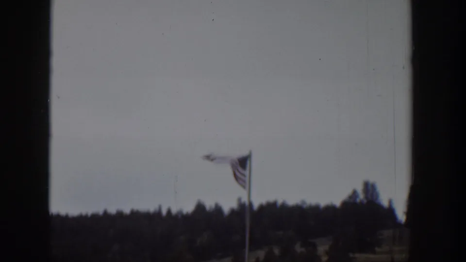 1939 Wyoming home-movie frame showing an American flag twisting in wind above a dark tree line and cloudy sky.