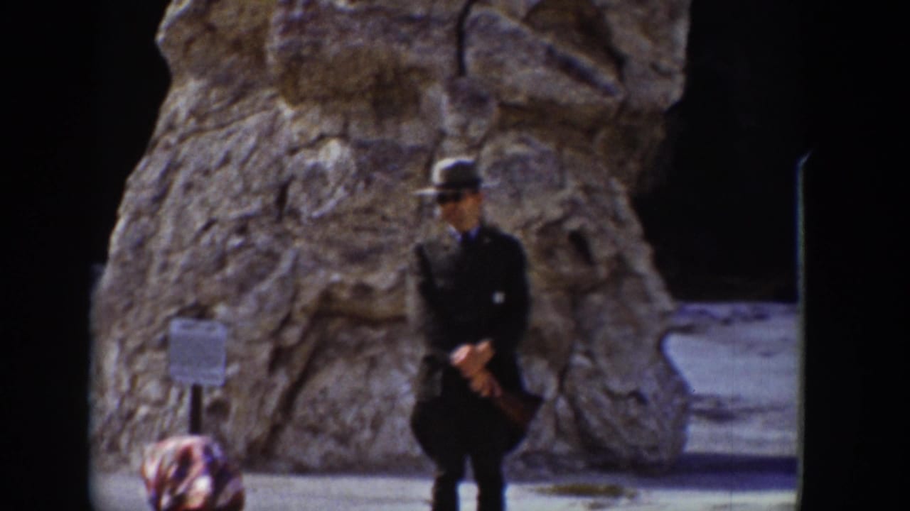 In this 1939 Wyoming home-movie frame, a man poses with hands clasped before a large weathered rock formation and a small sign.