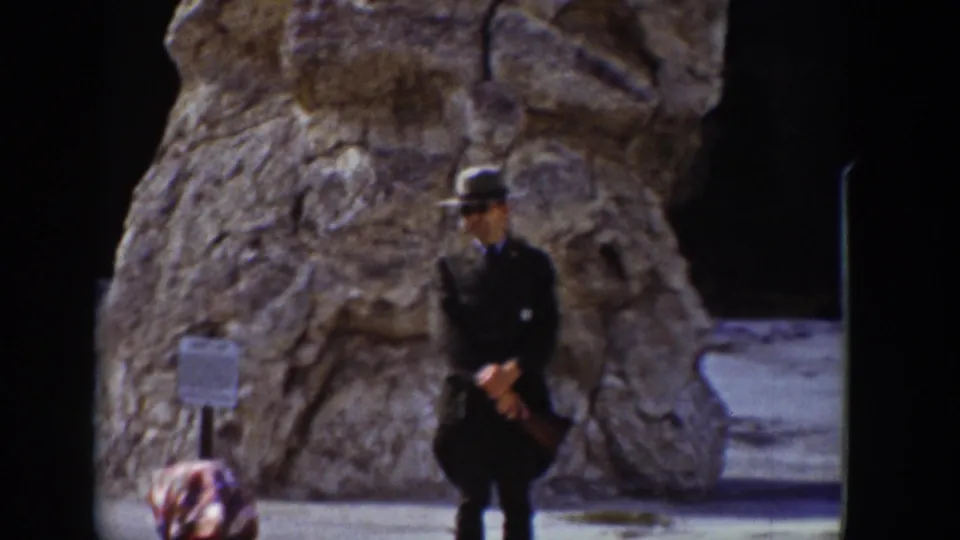 In this 1939 Wyoming home-movie frame, a man poses with hands clasped before a large weathered rock formation and a small sign.