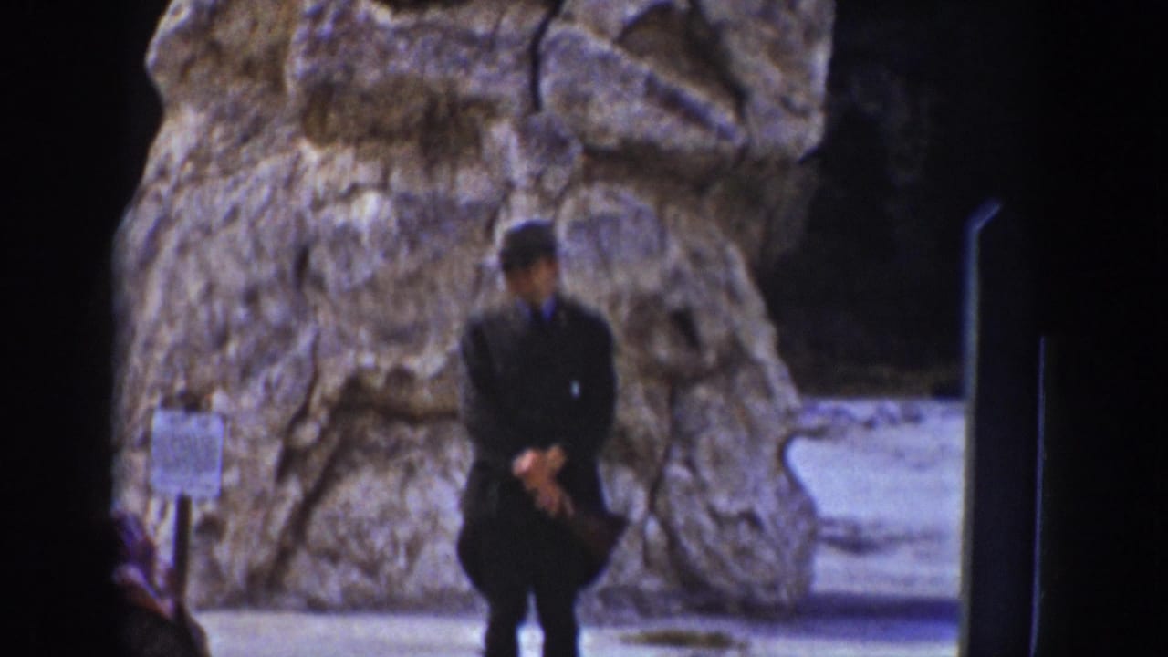 Man poses in front of a large rock formation with a sign nearby.