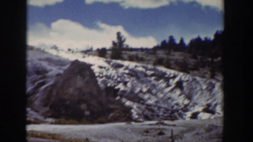 1939 Wyoming home-movie frame of a roadside slope with snow patches, trees, clouds, and a rock formation in the foreground.
