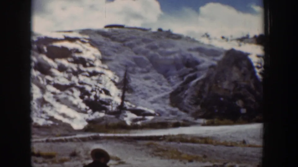 Wide Wyoming 8mm frame showing a snowy hillside, road, large rock, and a bundled person partly visible at the bottom edge.