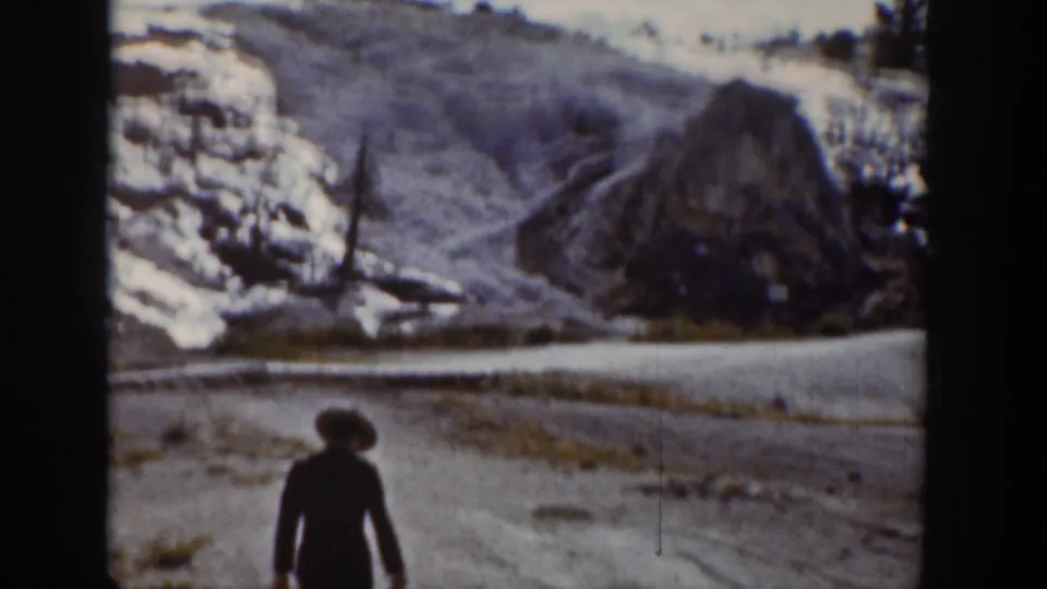 1939 Wyoming 8mm frame showing a woman in a dark coat and brimmed hat walking near a snow-lined mountain road.