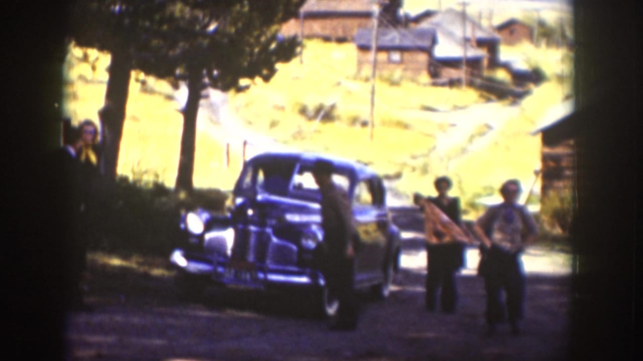 In 1939 Wyoming home-movie footage, adults gather beside a dark sedan on a dirt road near trees, cabins, and a grassy hillside.