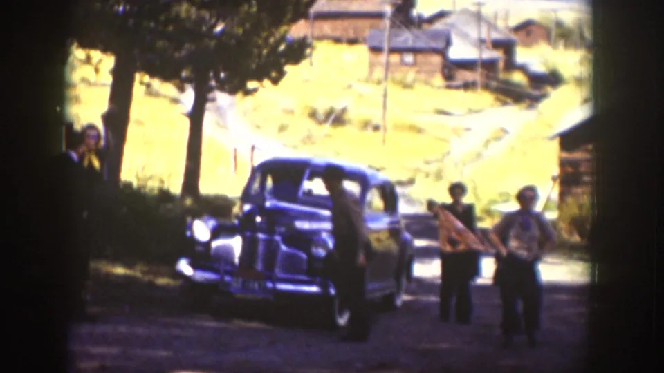 In 1939 Wyoming home-movie footage, adults gather beside a dark sedan on a dirt road near trees, cabins, and a grassy hillside.