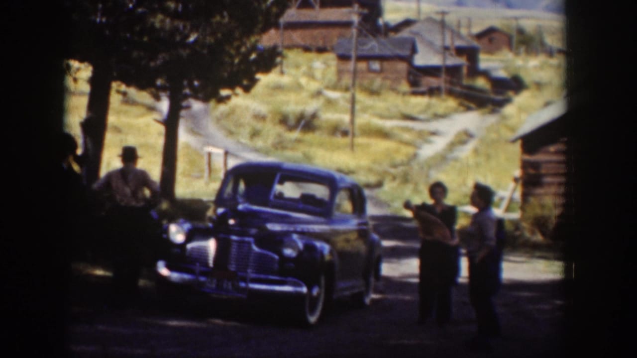 A dark sedan sits on a dirt road as several adults stand nearby under trees.