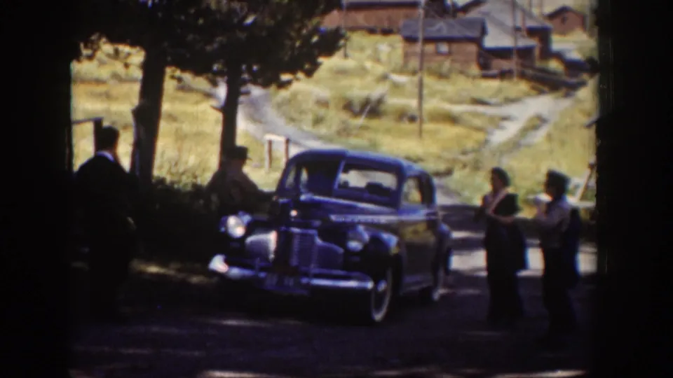 The home-movie frame shows adults near a parked sedan beneath trees, with a dirt lane, cabins, and dry hillside in the Wyoming background.