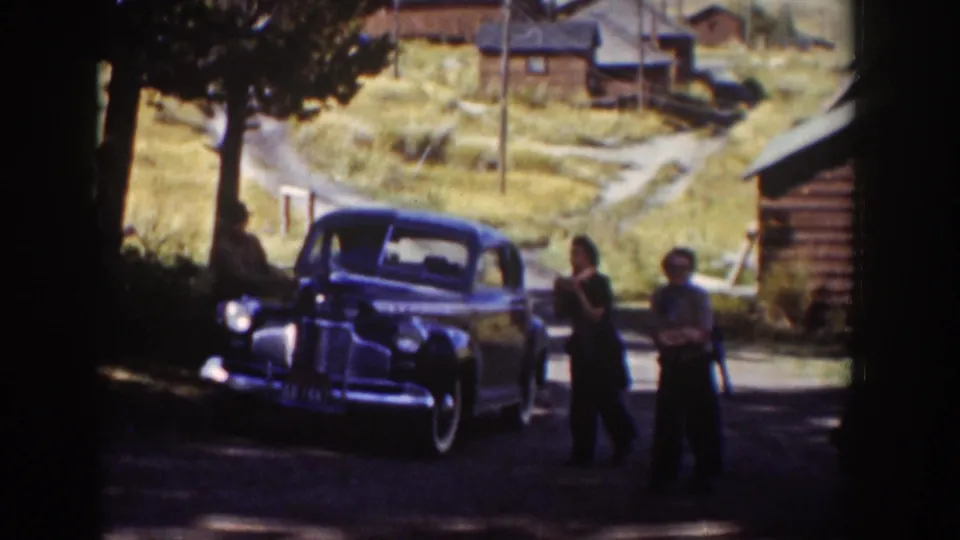 Two adults stand near a dark sedan on a Wyoming dirt road, framed by tree shade, log buildings, and a sunlit grassy hillside.