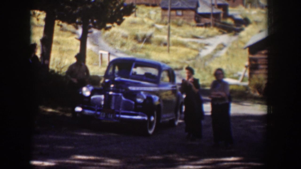 1939 Wyoming home-movie frame with adults near a dark blue sedan on a dirt road, with cabins and dry hillside grass in the background.
