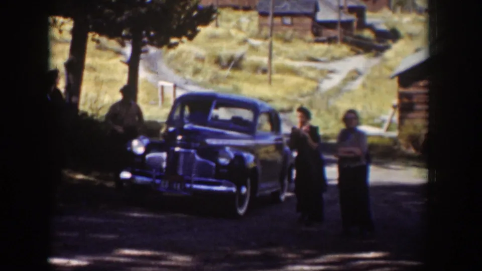 1939 Wyoming home-movie frame with adults near a dark blue sedan on a dirt road, with cabins and dry hillside grass in the background.