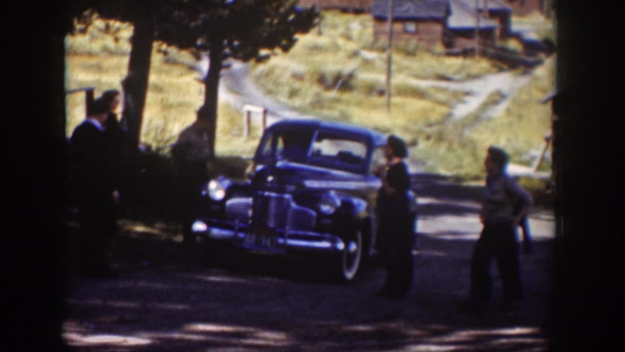 Adults in period clothing stand and move near a parked blue sedan in a rural Wyoming setting, with trees and cabins behind them.