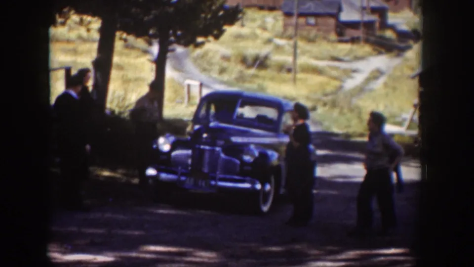 Adults in period clothing stand and move near a parked blue sedan in a rural Wyoming setting, with trees and cabins behind them.