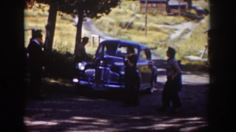 A group of adults gathers around a blue sedan on a shaded dirt road in the supplied 1939 Wyoming home-movie footage.