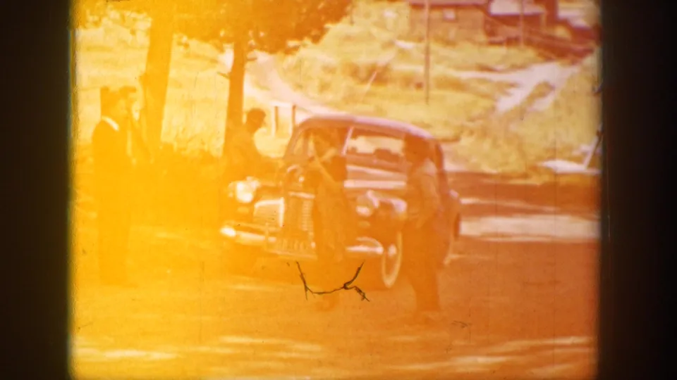 A strongly faded 8mm frame shows adults near the blue sedan on a Wyoming rural road, with orange exposure and visible film wear.