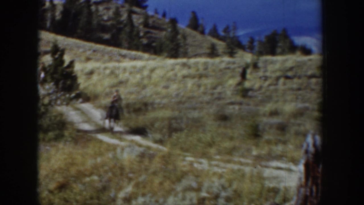 In 1939 Wyoming, a rider on horseback moves along a narrow path through dry grass below pines and rocky slopes.