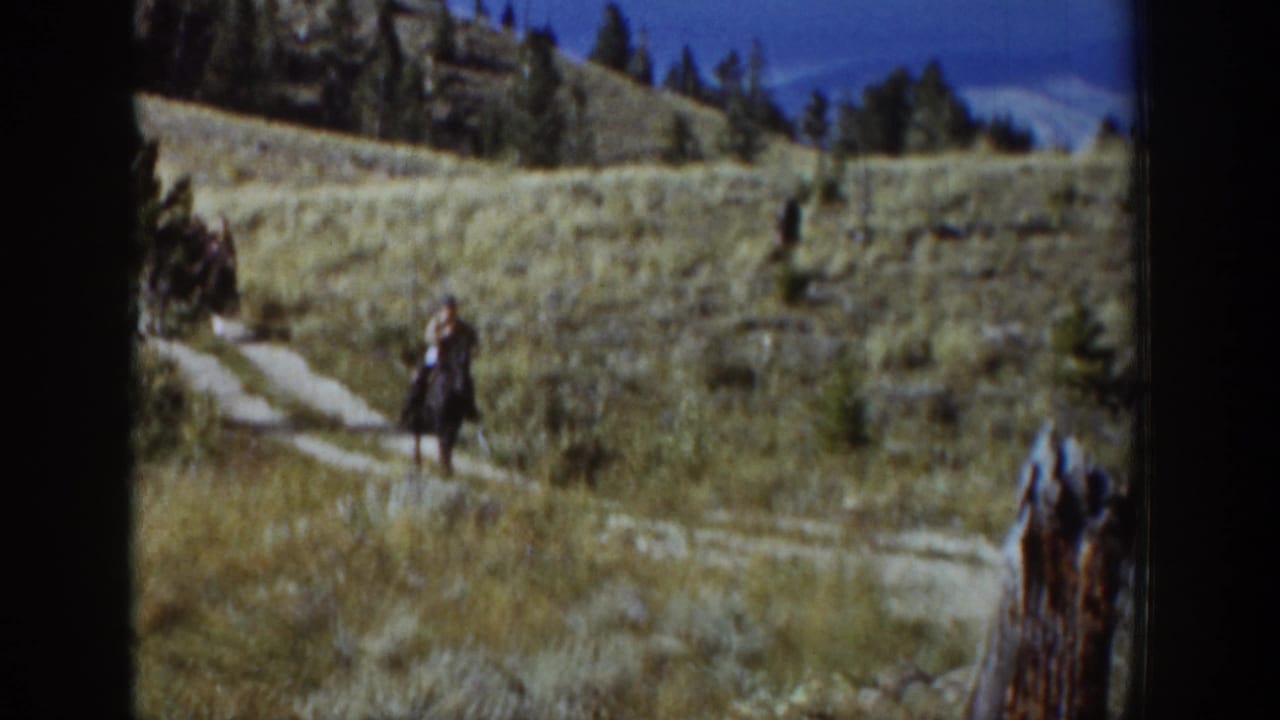 A horseback rider travels through Wyoming hill country, with dry grass, pines, rocks, and a partial horse at the frame edge.