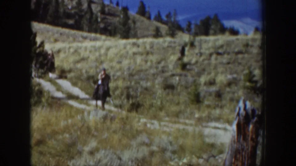 A horseback rider travels through Wyoming hill country, with dry grass, pines, rocks, and a partial horse at the frame edge.