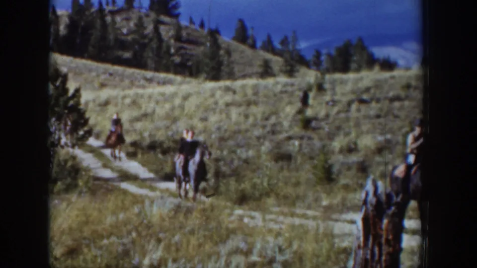 A small group of riders on horseback appears on a Wyoming hillside trail, with trees and rocky ground in the background.
