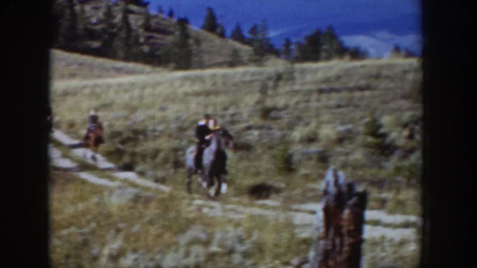 Riders on horseback travel across a 1939 Wyoming hillside, with a winding dirt path, dry grass, and pines behind them.