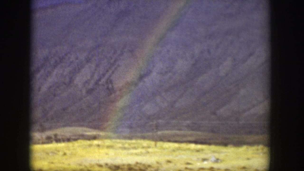 1939 Wyoming 8mm frame showing a rainbow slanting over rugged mountains and a pale valley floor beneath heavy cloud cover.