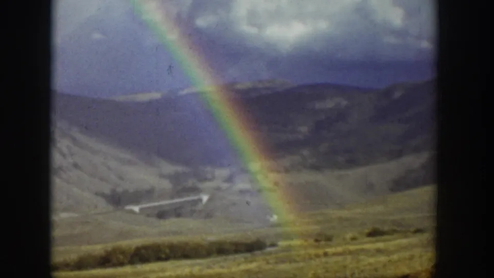 1939 Wyoming home-movie frame with a vivid rainbow descending across hills, open valley land, and distant mountain ridges.