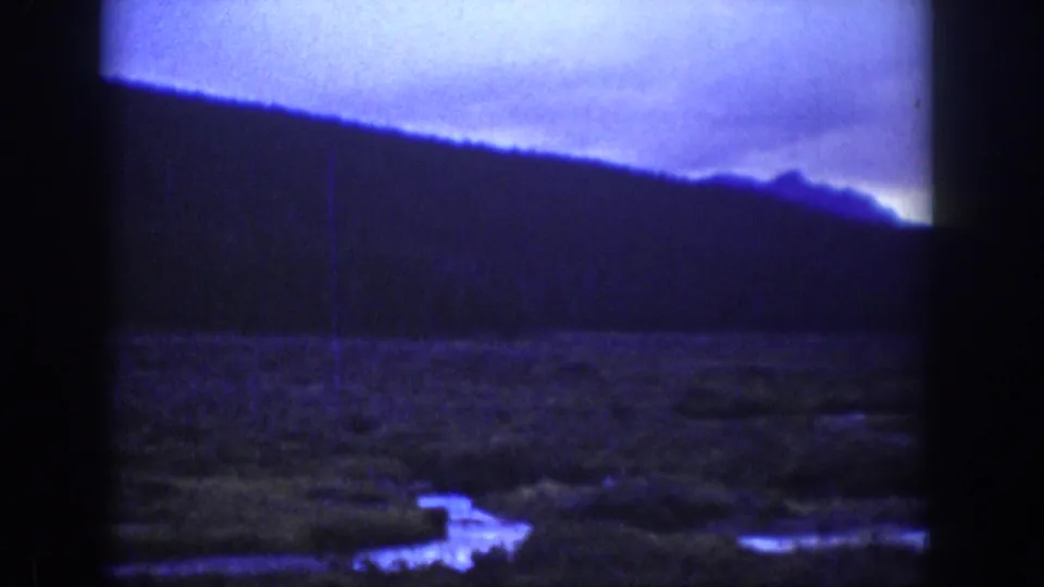 Wyoming 1939 home-movie frame showing dark wooded hills, cloudy sky, and reflective lowland water with strong blue-purple film tint.